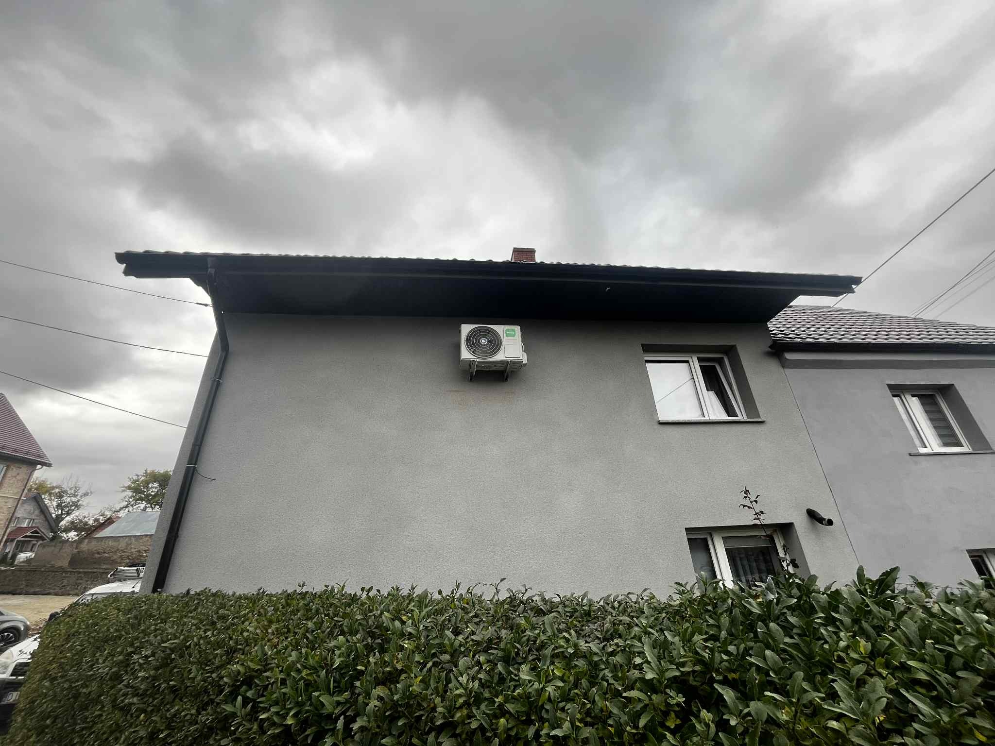 Szary dom z zamontowaną klimatyzacją na ścianie, widok z dołu, pochmurne niebo w tle. Krzewy przed budynkiem.