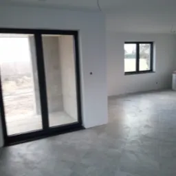 Wnętrze w stanie deweloperskim z szarą, matową podłogą z płytek, dużymi przesuwnymi drzwiami balkonowymi z czarną ramą i widokiem na zewnątrz oraz oknem z czarną ramą po prawej stronie