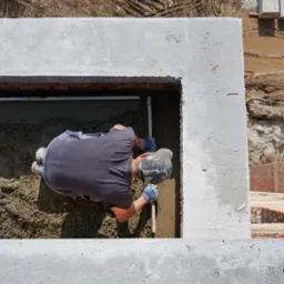 Pracownik w czapce moro wyrównuje poziom wylewki wewnątrz betonowej ramy, używając poziomicy i łaty; widok z góry.