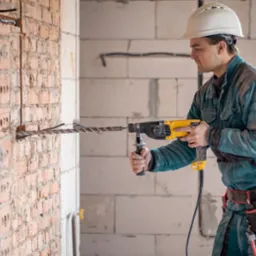 Mężczyzna w kasku wierci otwór w ceglanej ścianie za pomocą wiertarki udarowej z długim wiertłem spiralnym, widoczne narzędzia w pasie monterskim.
