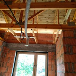 Recupero - Wnętrze budynku w trakcie instalacji rekuperacji: widoczny surowy strop drewniany, ściany z czerwonej cegły i rura wentylacyjna podwieszona do belek stropowych, okno w tle.