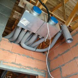 Recupero - Centrala wentylacyjna z rekuperacją, widoczna na tle surowej konstrukcji dachu i muru z cegieł kratówek, z podłączonymi przewodami wentylacyjnymi, niebieskie pokrywy.