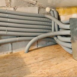 Recupero - Szereg szarych, karbowanych rur wentylacyjnych przymocowanych do ściany z betonowych pustaków, prowadzących do metalowej skrzynki z okrągłymi wlotami, zamontowanej na podłodze z płyt OSB.