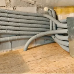 Szereg szarych, karbowanych rur wentylacyjnych przymocowanych do ściany z betonowych pustaków, prowadzących do metalowej skrzynki z okrągłymi wlotami, zamontowanej na podłodze z płyt OSB.