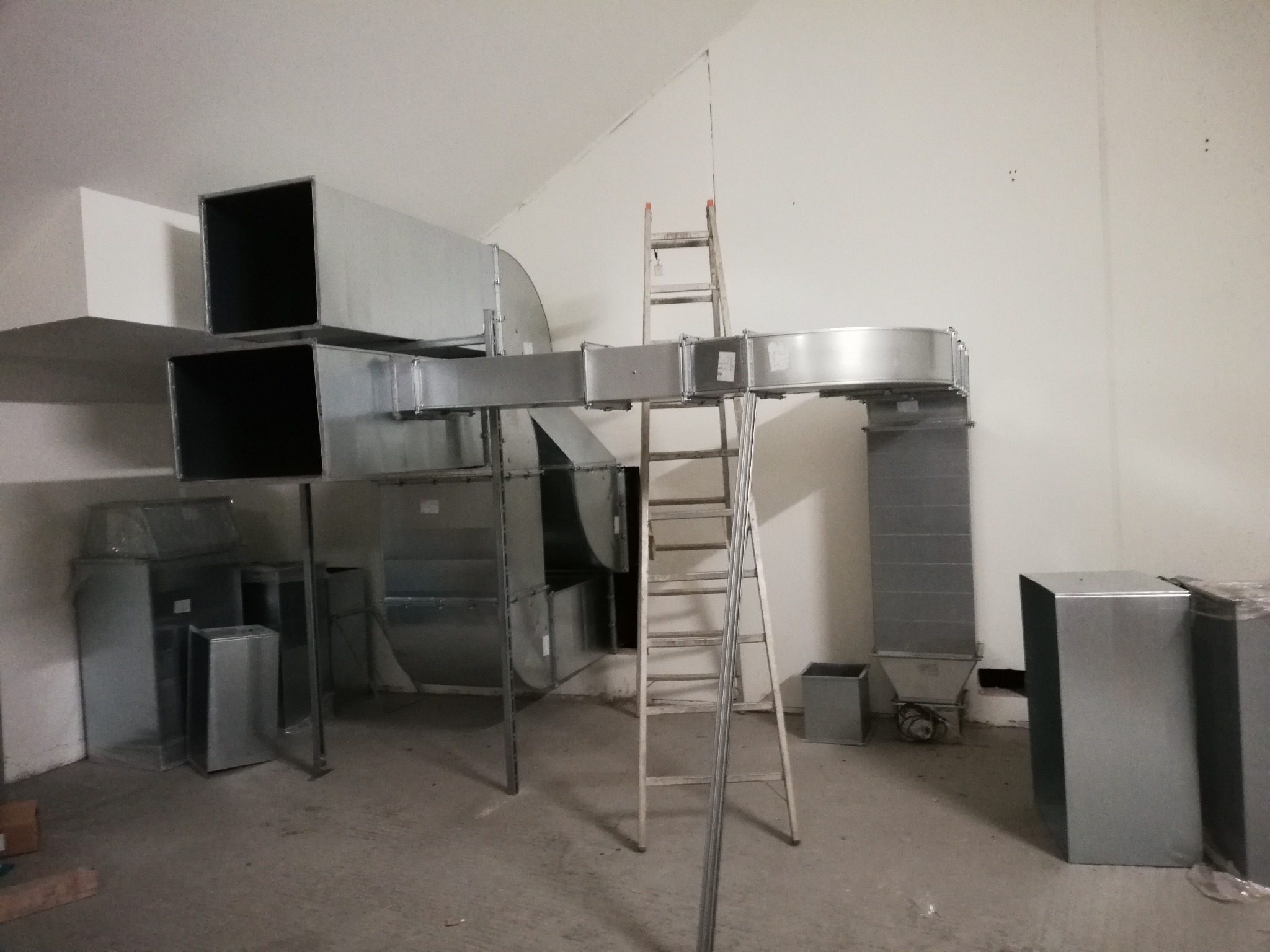 Montaż systemu wentylacji z metalowych kanałów, widoczna drabina oparta o ścianę, surowy stan wykończenia wnętrza.