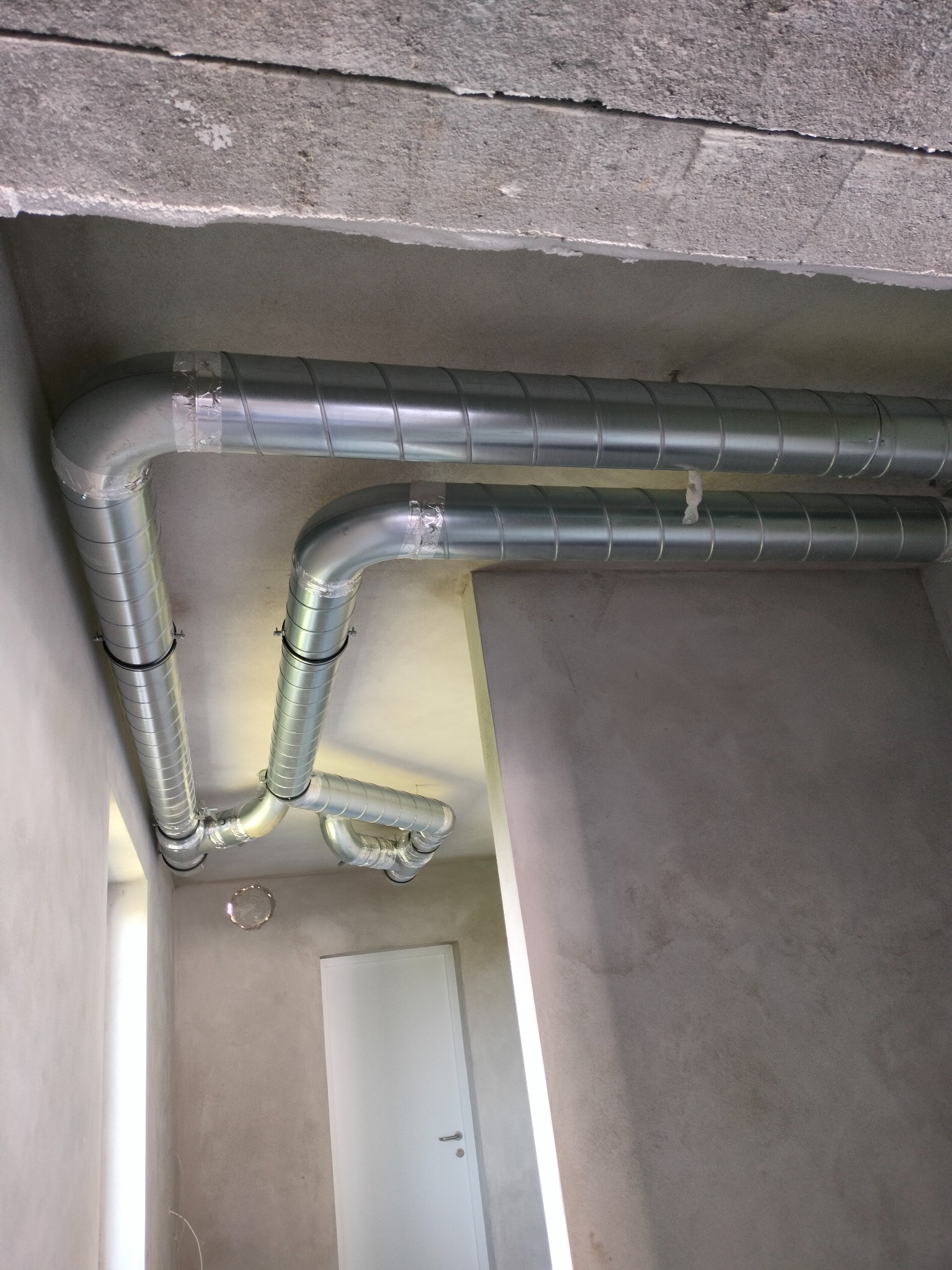 Spiralne, metalowe kanały wentylacyjne zamontowane pod surowym, betonowym stropem, zabezpieczone taśmą i obejmami, widoczne w surowym wnętrzu z białą ścianą i drzwiami.