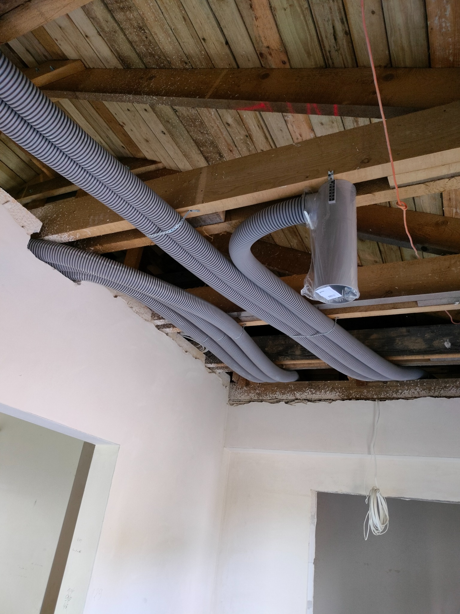 Szare, karbowane rury wentylacyjne prowadzone pod drewnianym stropem, widoczny fragment instalacji z filtrem w folii, surowy stan wykończenia wnętrza.