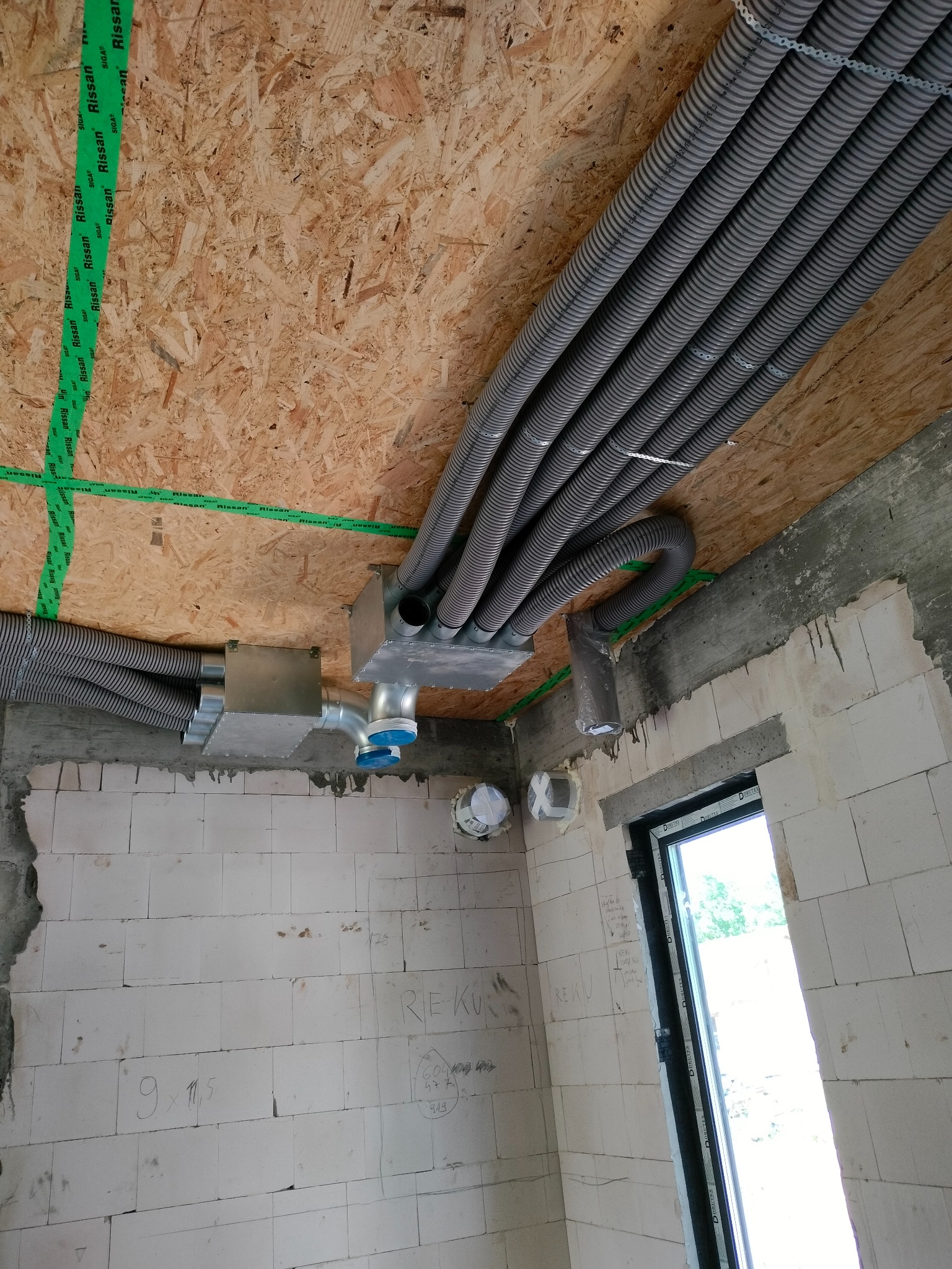 Wnętrze w trakcie montażu instalacji wentylacyjnej; widoczne szare, karbowane rury wentylacyjne przymocowane do drewnianego sufitu z zieloną taśmą uszczelniającą, metalowe skrzynki rozdzielcze...