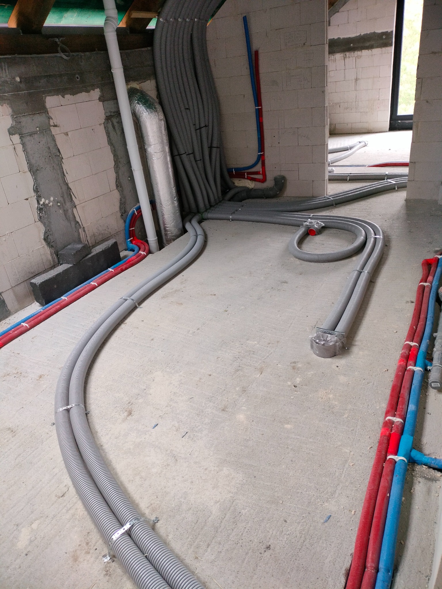 Szare, karbowane przewody wentylacyjne prowadzone po surowej betonowej podłodze w budynku w trakcie budowy, z widocznymi czerwonymi i niebieskimi rurami instalacyjnymi przy ścianach z bloków.