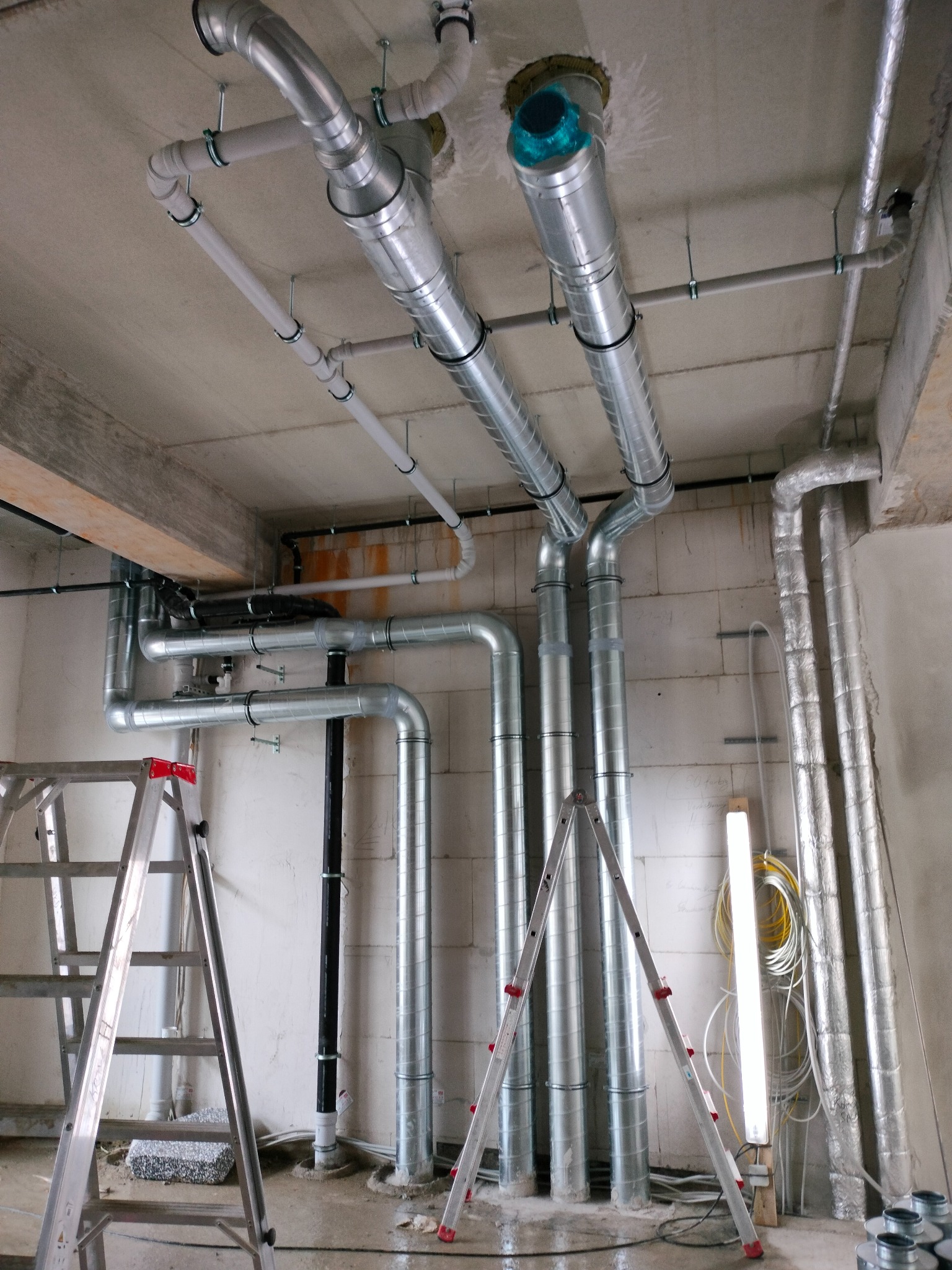 Wnętrze w trakcie montażu systemu wentylacji: widoczne rury wentylacyjne ze stali ocynkowanej, prowadzone pod sufitem i wzdłuż ściany z betonu komórkowego, dwie rozstawione aluminiowe drabiny...