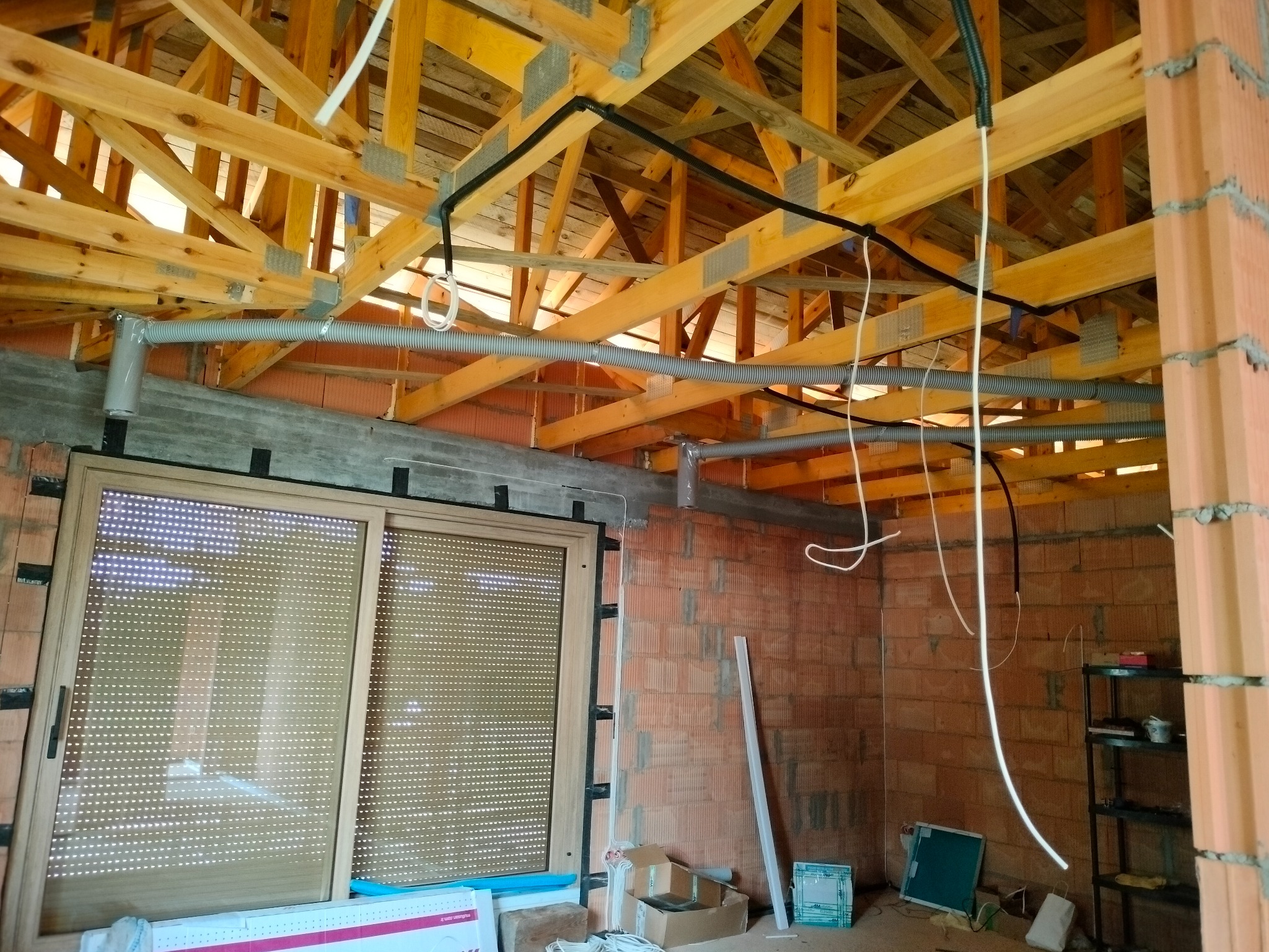 Wnętrze w trakcie budowy z widoczną instalacją wentylacyjną pod drewnianą konstrukcją dachu, szara rura wentylacyjna biegnie wzdłuż ściany z cegły, okno z roletami w tle.