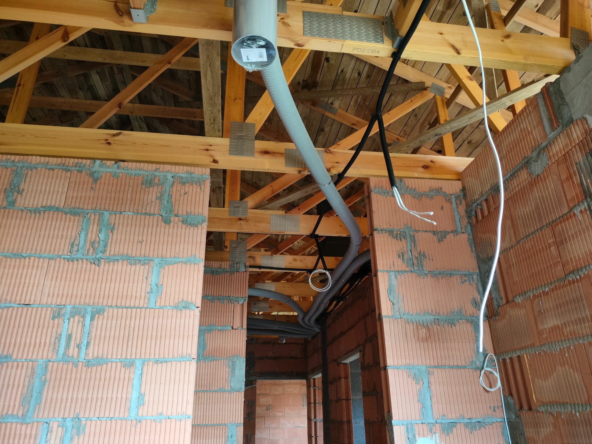 Wnętrze budynku w trakcie budowy z widocznymi przewodami wentylacyjnymi i kablami elektrycznymi pod drewnianą konstrukcją dachu.