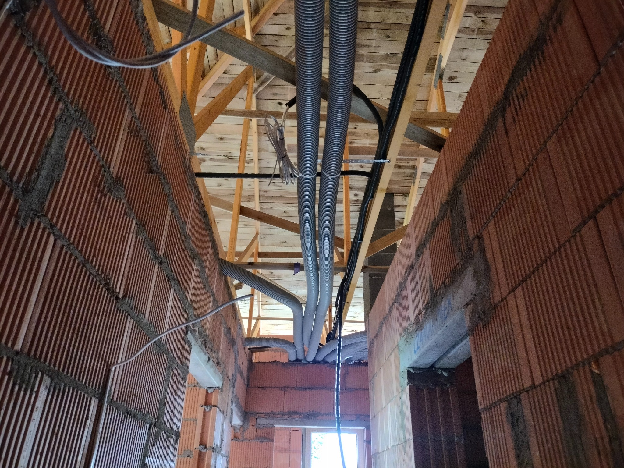 Wnętrze budynku w trakcie budowy z widocznymi rurami wentylacyjnymi z tworzywa sztucznego prowadzonymi pod drewnianą konstrukcją dachu, ściany z czerwonej cegły dziurawki.