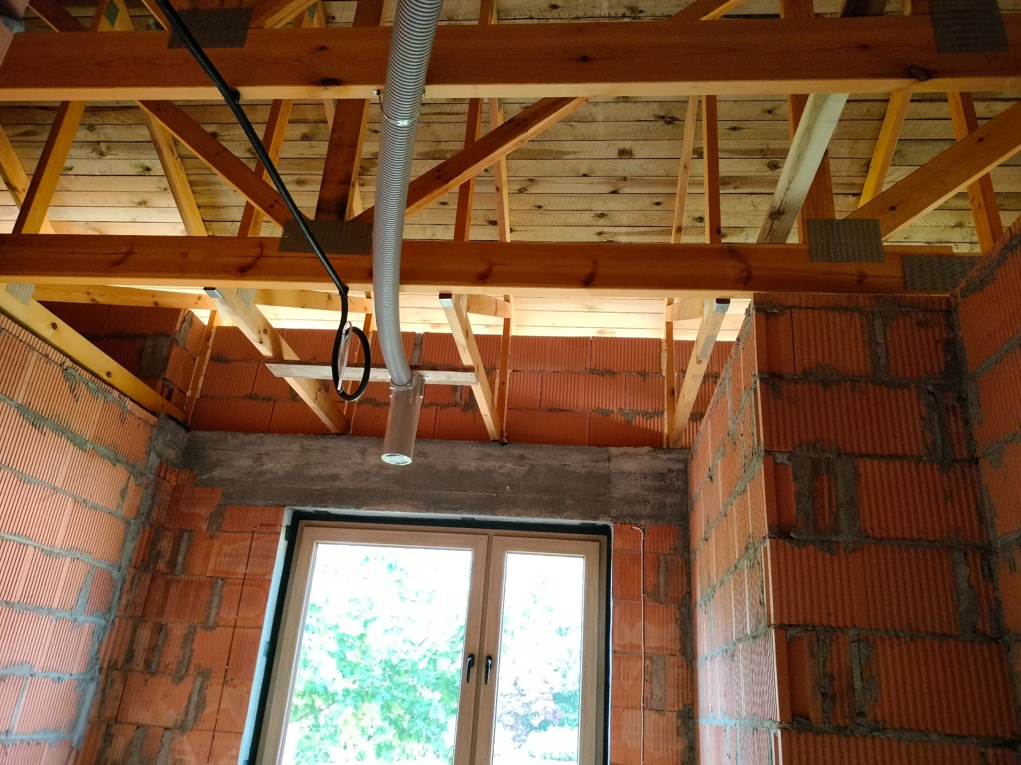 Wnętrze budynku w trakcie instalacji rekuperacji: widoczny surowy strop drewniany, ściany z czerwonej cegły i rura wentylacyjna podwieszona do belek stropowych, okno w tle.