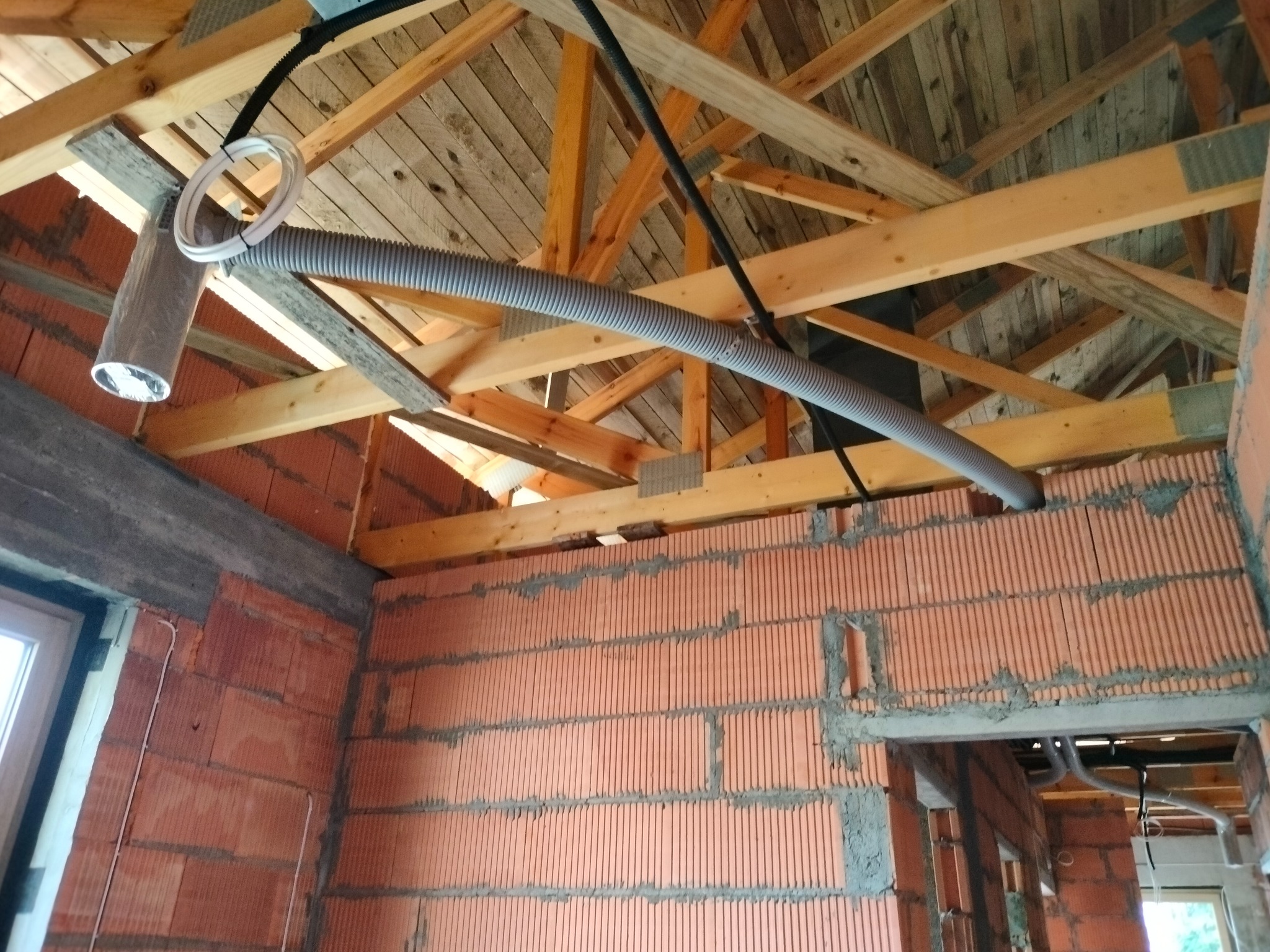 Szary, elastyczny przewód wentylacyjny podwieszony pod drewnianą konstrukcją dachu w surowym, ceglanym budynku w trakcie budowy. Widoczne elementy montażowe i nieocieplony kanał wentylacyjny.