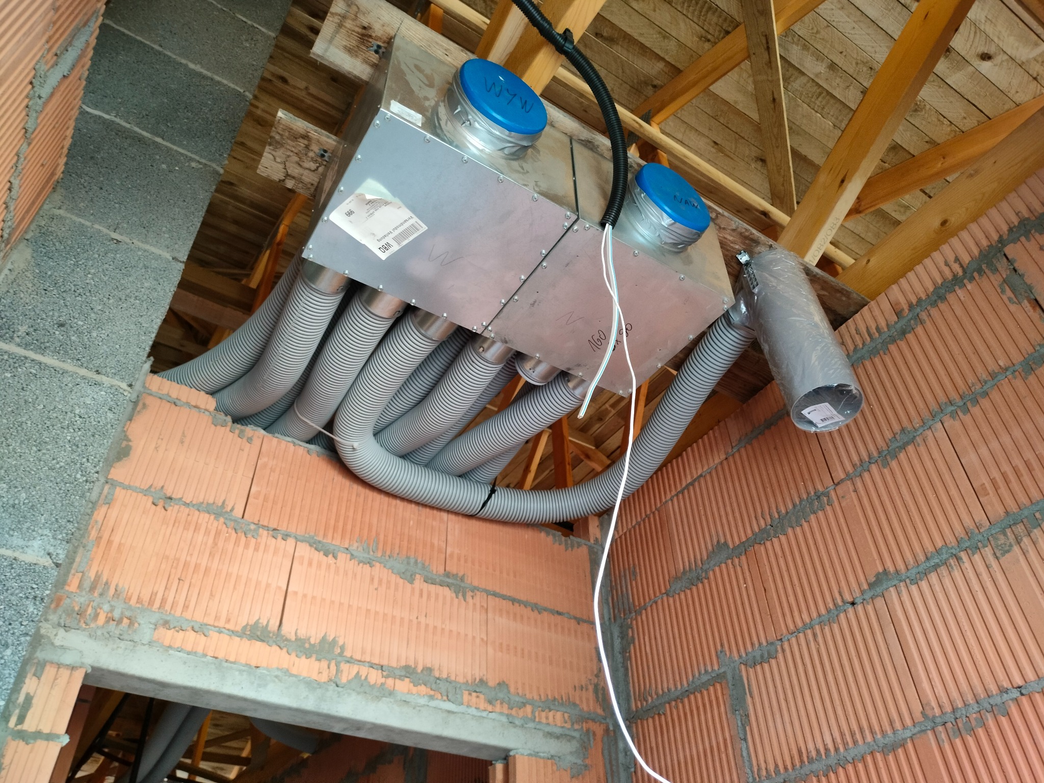 Centrala wentylacyjna z rekuperacją, widoczna na tle surowej konstrukcji dachu i muru z cegieł kratówek, z podłączonymi przewodami wentylacyjnymi, niebieskie pokrywy.