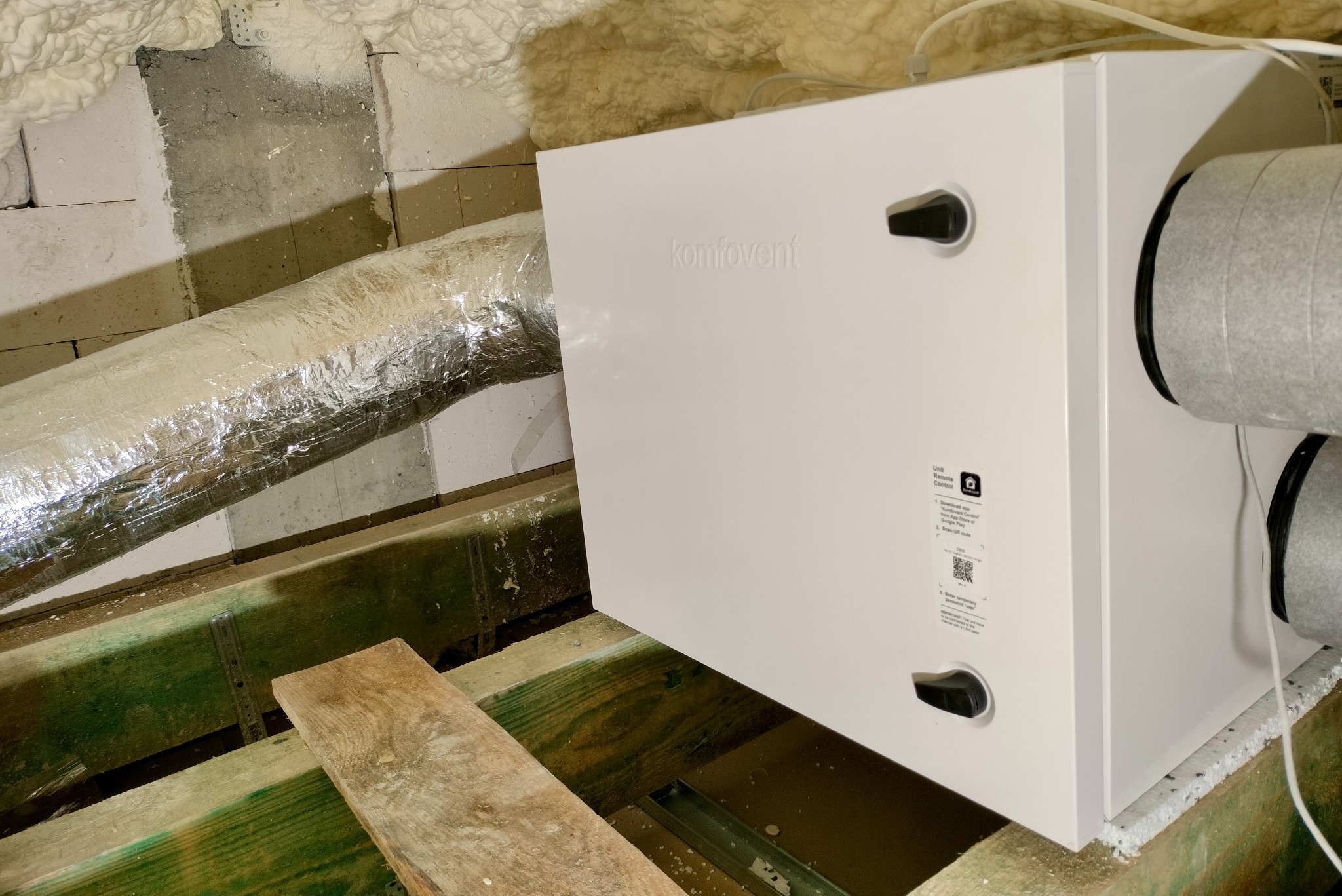 Biała jednostka rekuperacyjna Komfovent z podłączonymi przewodami wentylacyjnymi, zamontowana na drewnianej konstrukcji pod stropem z izolacją z pianki.
