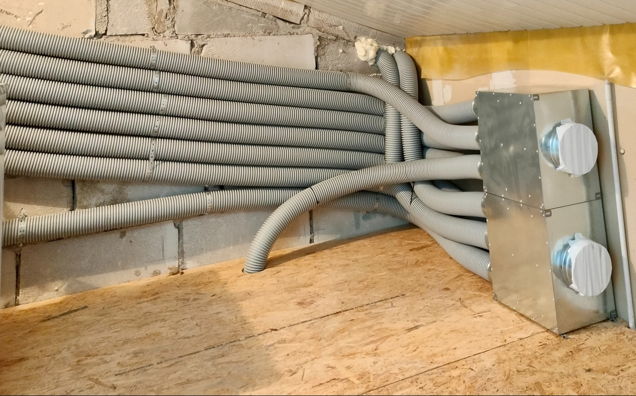 Szereg szarych, karbowanych rur wentylacyjnych przymocowanych do ściany z betonowych pustaków, prowadzących do metalowej skrzynki z okrągłymi wlotami, zamontowanej na podłodze z płyt OSB.