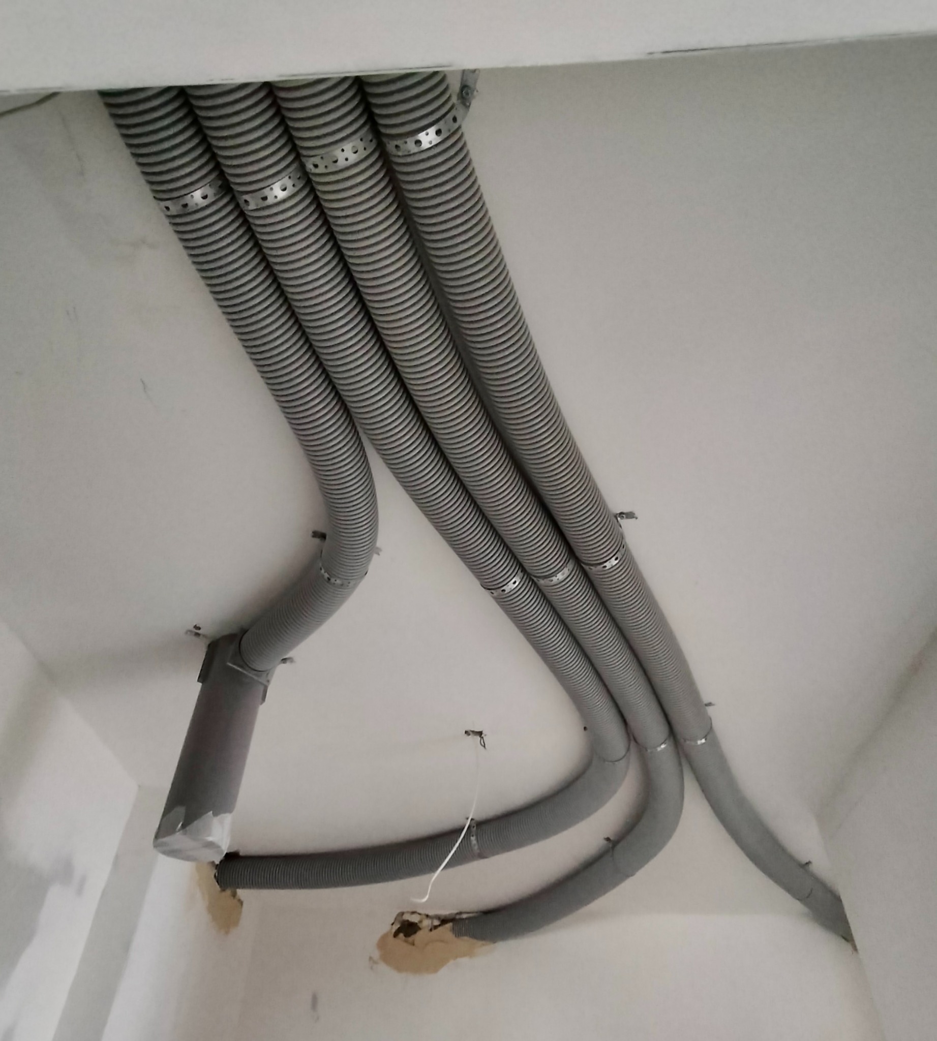 Szare, elastyczne rury wentylacyjne przymocowane do sufitu za pomocą metalowych obejm, widoczne niedoskonałości wykończenia ścian i sufitu.
