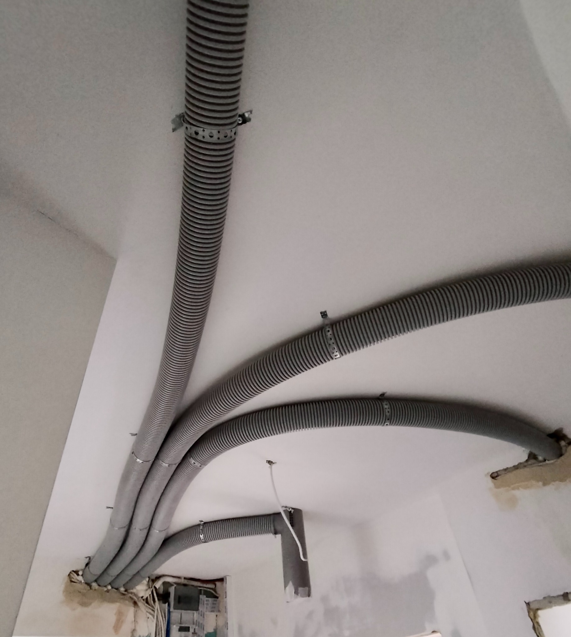 Szare, karbowane rury wentylacyjne przymocowane do sufitu za pomocą metalowych obejm, widoczne niedokończone wykończenie ścian i otwory w suficie.