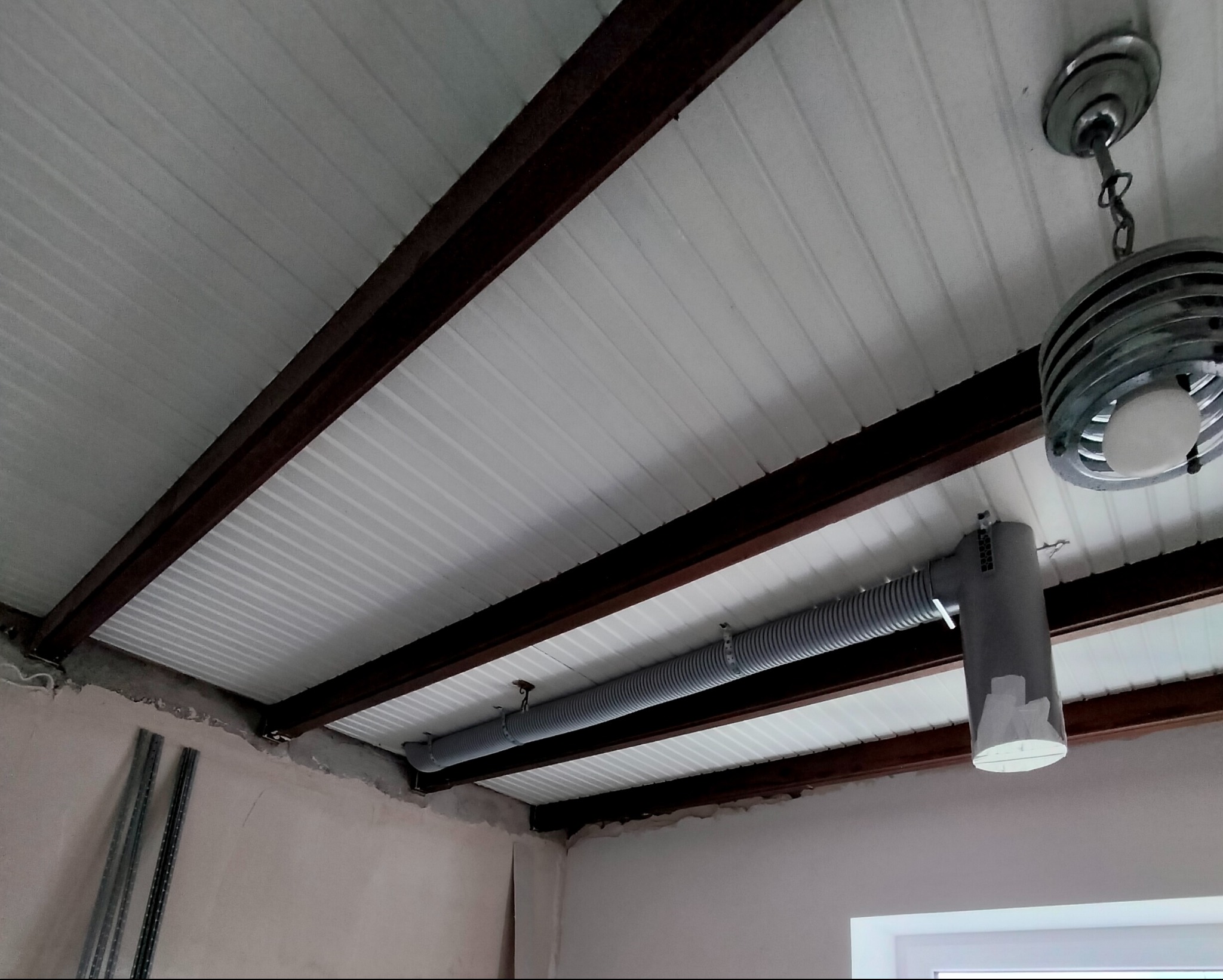 Szary, elastyczny przewód wentylacyjny zamontowany pod białym sufitem panelowym, podtrzymywany przez brązowe, metalowe belki. Widoczna lampa wisząca w tle oraz fragmenty ściany w stanie surowym...