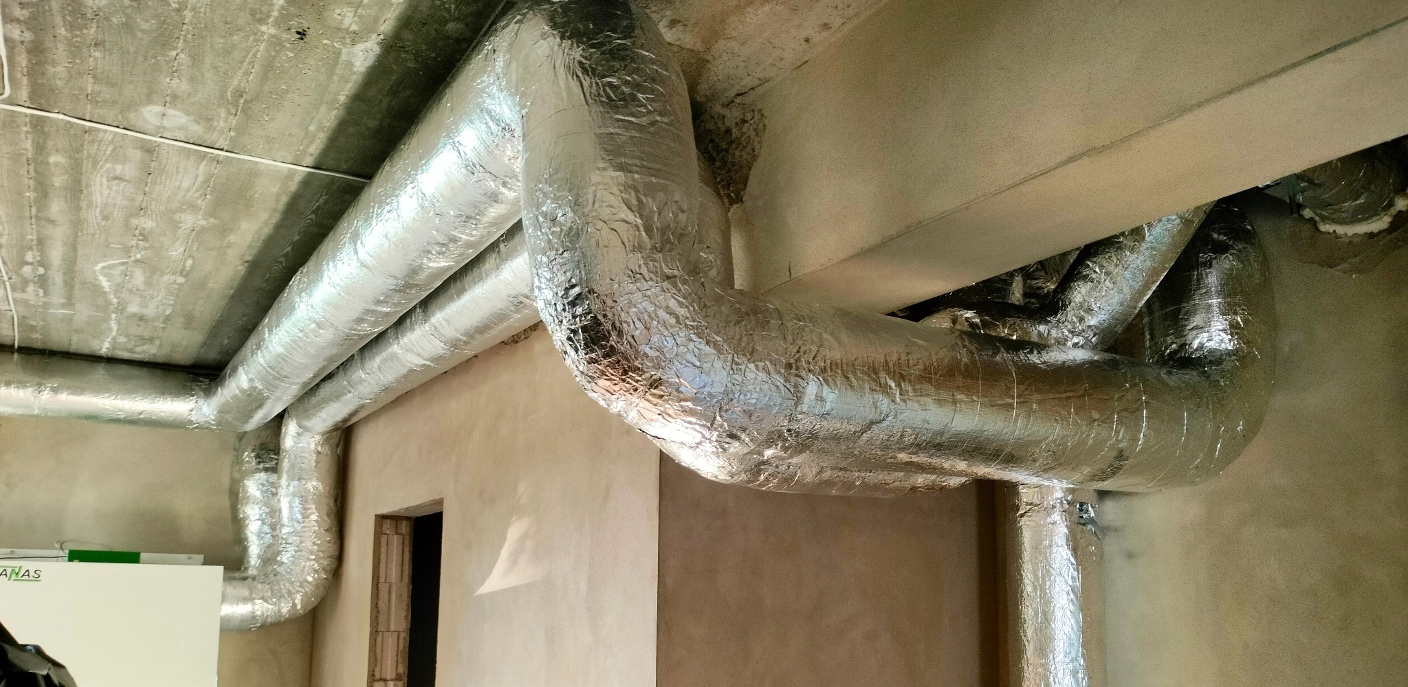 Srebrne, zaizolowane rury wentylacyjne pod betonowym sufitem w surowym wnętrzu budynku, widoczny fragment urządzenia wentylacyjnego i otwór drzwiowy.