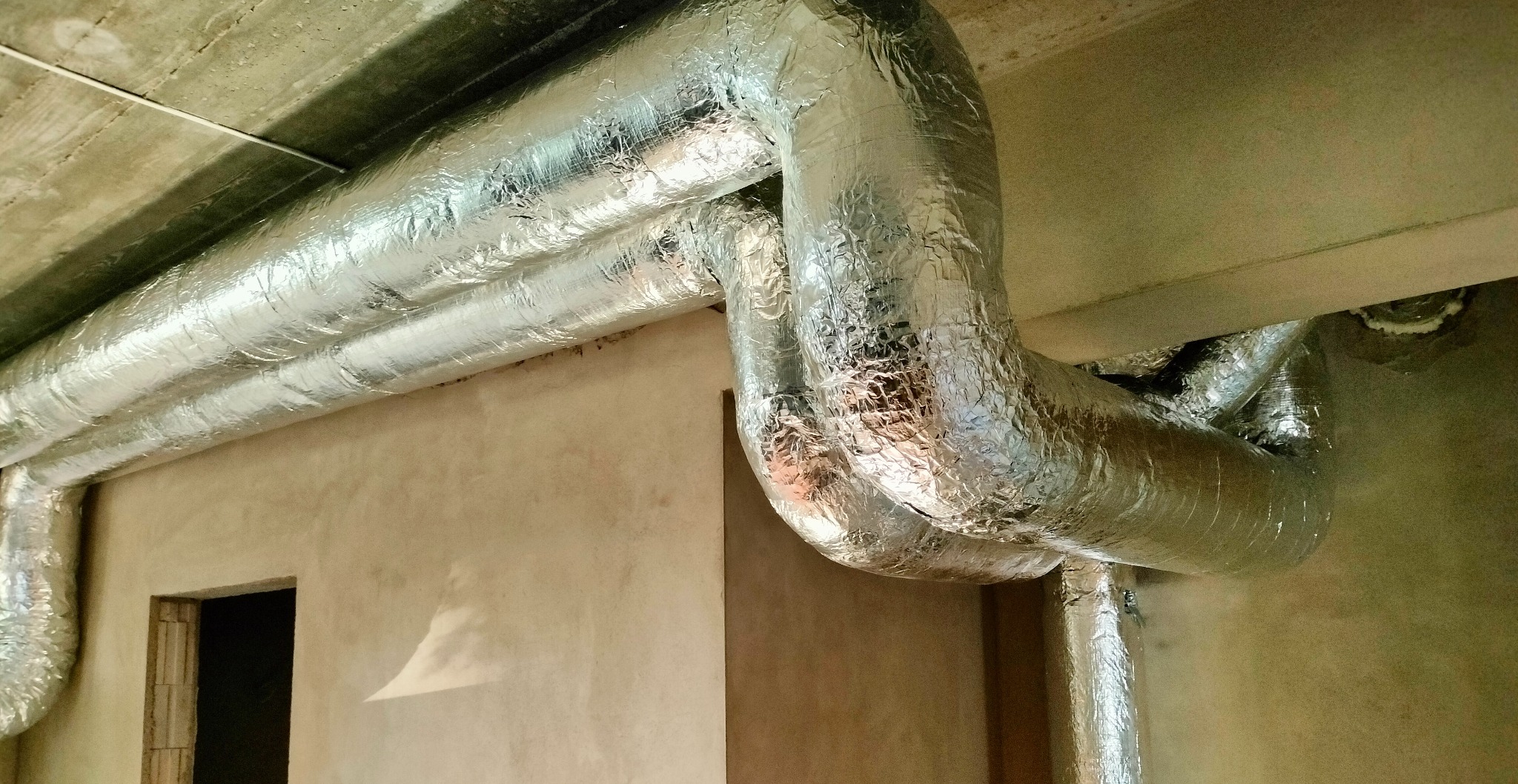 Izolowane, srebrne rury wentylacyjne biegnące pod betonowym sufitem w surowym wnętrzu budynku, widoczny otwór drzwiowy w ścianie.