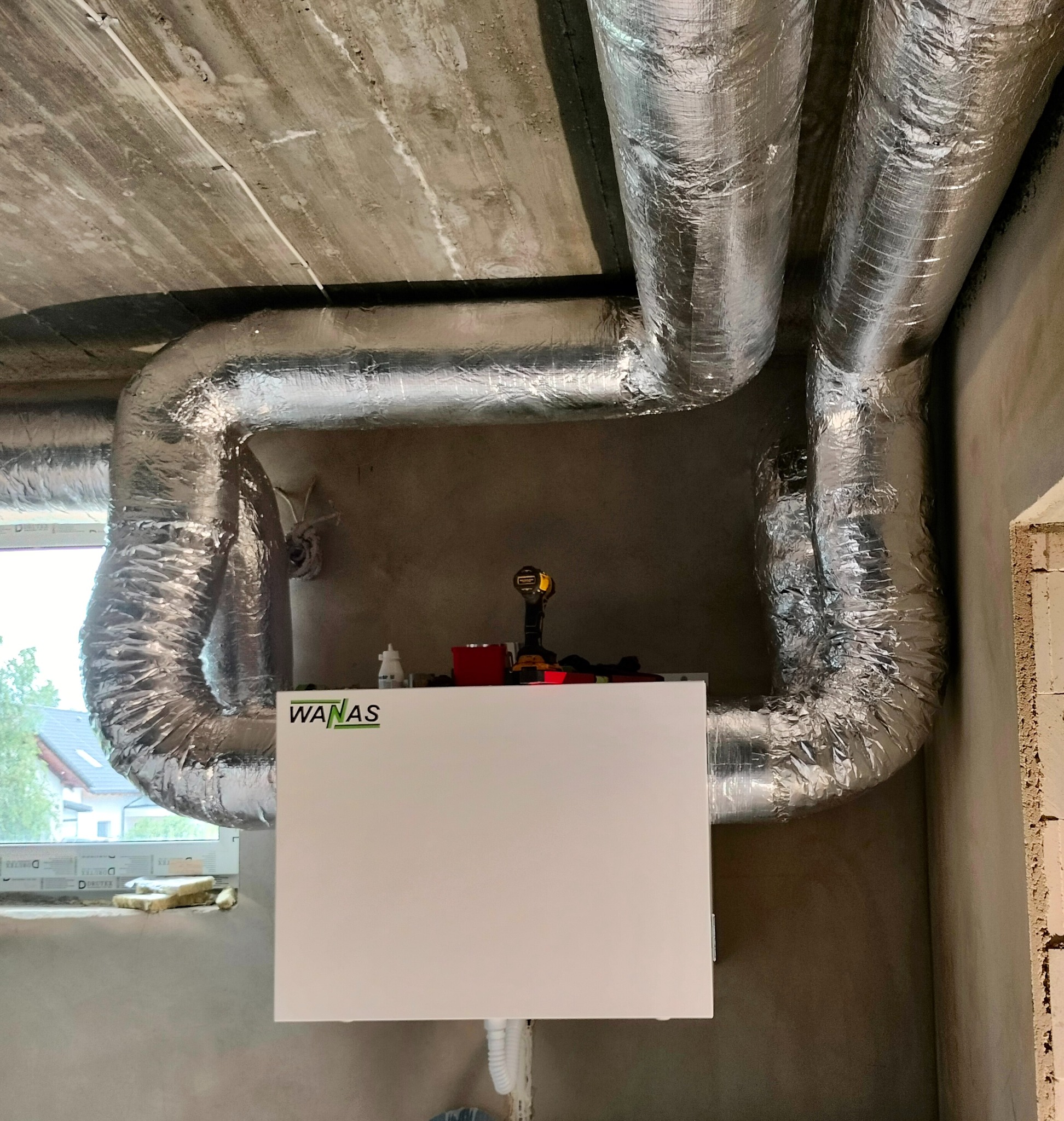 Biała centrala wentylacyjna WANAS z izolowanymi przewodami wentylacyjnymi w surowym wnętrzu budynku w trakcie montażu, widoczna wiertarka akumulatorowa i narzędzia na obudowie centrali.