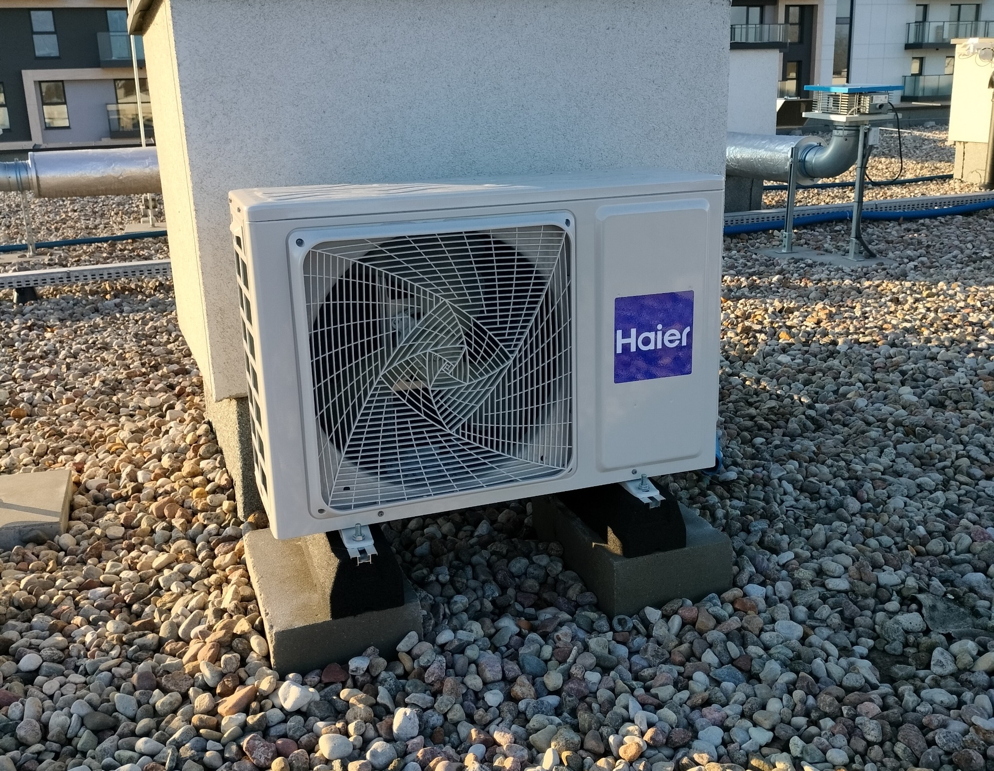 Zewnętrzna jednostka klimatyzacji marki Haier zamontowana na betonowych podstawach na żwirowym dachu budynku.