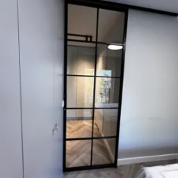 Przesuwne drzwi wewnętrzne w stylu loft z czarnymi szprosami, widoczne z boku, z elementami szafy w tle i parkietem w jodełkę.