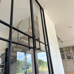 PRO-SPAW Bartosz Białas - Szklana ściana działowa z czarnymi metalowymi profilami, widoczne schody drewniane z metalową balustradą oraz regał z książkami w tle.