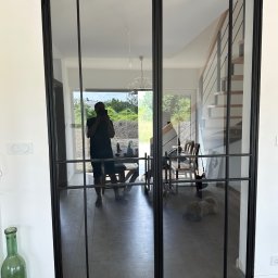 PRO-SPAW Bartosz Białas - Dwuskrzydłowe drzwi wewnętrzne ze szprosami w czarnej ramie, widoczne schody z metalową balustradą i drewniane stopnie, odbicie osoby robiącej zdjęcie, butelka w rogu.