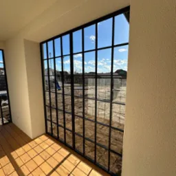 Duże, przeszklone okno z czarnymi szprosami, widok z wnętrza na plac budowy z domami w tle, drewniana podłoga tarasowa w jasnym odcieniu.