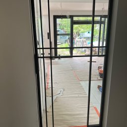 PRO-SPAW Bartosz Białas - Czarna, metalowa rama drzwiowa z szybami, widoczna w trakcie montażu w nowym domu, z zabezpieczonymi podłogami i widoczną osobą oraz narzędziami w tle.