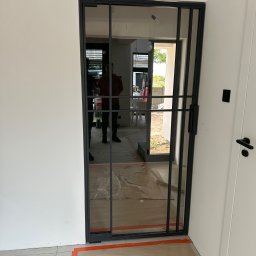 PRO-SPAW Bartosz Białas - Szklane drzwi przesuwne z czarną metalową ramą, widoczne zabezpieczenia podłogi i odbicie osoby wewnątrz budynku.