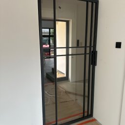 PRO-SPAW Bartosz Białas - Nowoczesne, przesuwne drzwi wewnętrzne z czarnymi metalowymi szprosami i szybami, widok na wnętrze domu z betonowymi schodami i oknem, zabezpieczone kartonem i taśmą malarską.