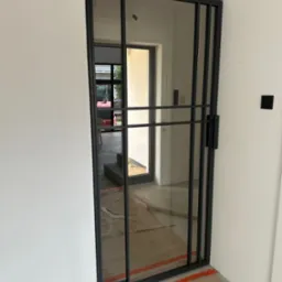 Nowoczesne, przesuwne drzwi wewnętrzne z czarnymi metalowymi szprosami i szybami, widok na wnętrze domu z betonowymi schodami i oknem, zabezpieczone kartonem i taśmą malarską.