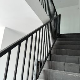 PRO-SPAW Bartosz Białas - Wewnętrzne schody wyłożone szarymi płytkami, z czarną metalową balustradą o pionowych szczeblach, widoczne przewody elektryczne na suficie.