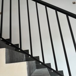 PRO-SPAW Bartosz Białas - Schody z czarną metalową balustradą o prostych szczeblach, wykończone płytkami imitującymi kamień, widoczne niedoskonałości malowania ścian.