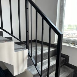 PRO-SPAW Bartosz Białas - Nowoczesne schody z czarną, metalową balustradą o prostym wzorze, widok od dołu, częściowo wykończone, z widocznym oknem w tle.