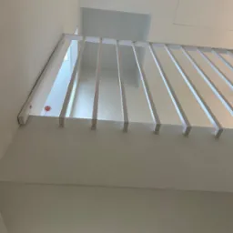 Minimalistyczna, biała balustrada z pionowych prętów, widok z dołu na jasnym tle ściany i sufitu, z widocznym fragmentem otworu w stropie.