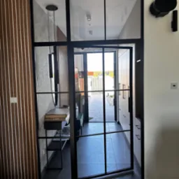 Nowoczesne, czarne, przeszklone drzwi wewnętrzne z metalową ramą, widoczne z perspektywy wnętrza domu, z elementami dekoracyjnymi i oświetleniem w tle.