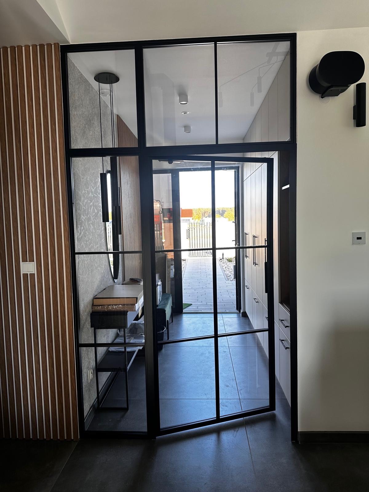 Nowoczesne, czarne, przeszklone drzwi wewnętrzne z metalową ramą, widoczne z perspektywy wnętrza domu, z elementami dekoracyjnymi i oświetleniem w tle.