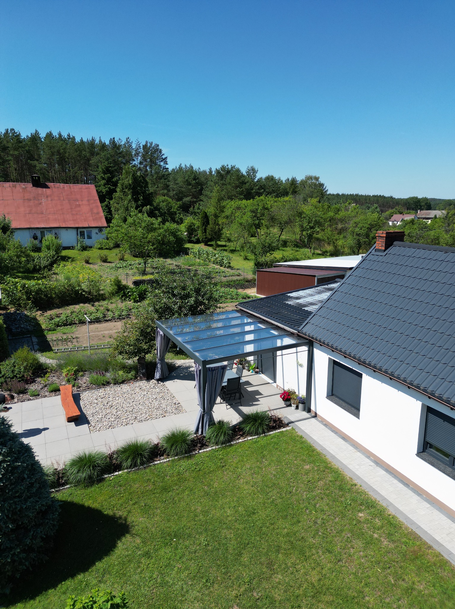 Nowoczesna pergola z szarymi zasłonami i szklanym dachem przylegająca do białego domu z szarą dachówką, widok z góry na zadbany ogród z trawnikiem, kamienną ścieżką, rabatami i drewnianą ławką.