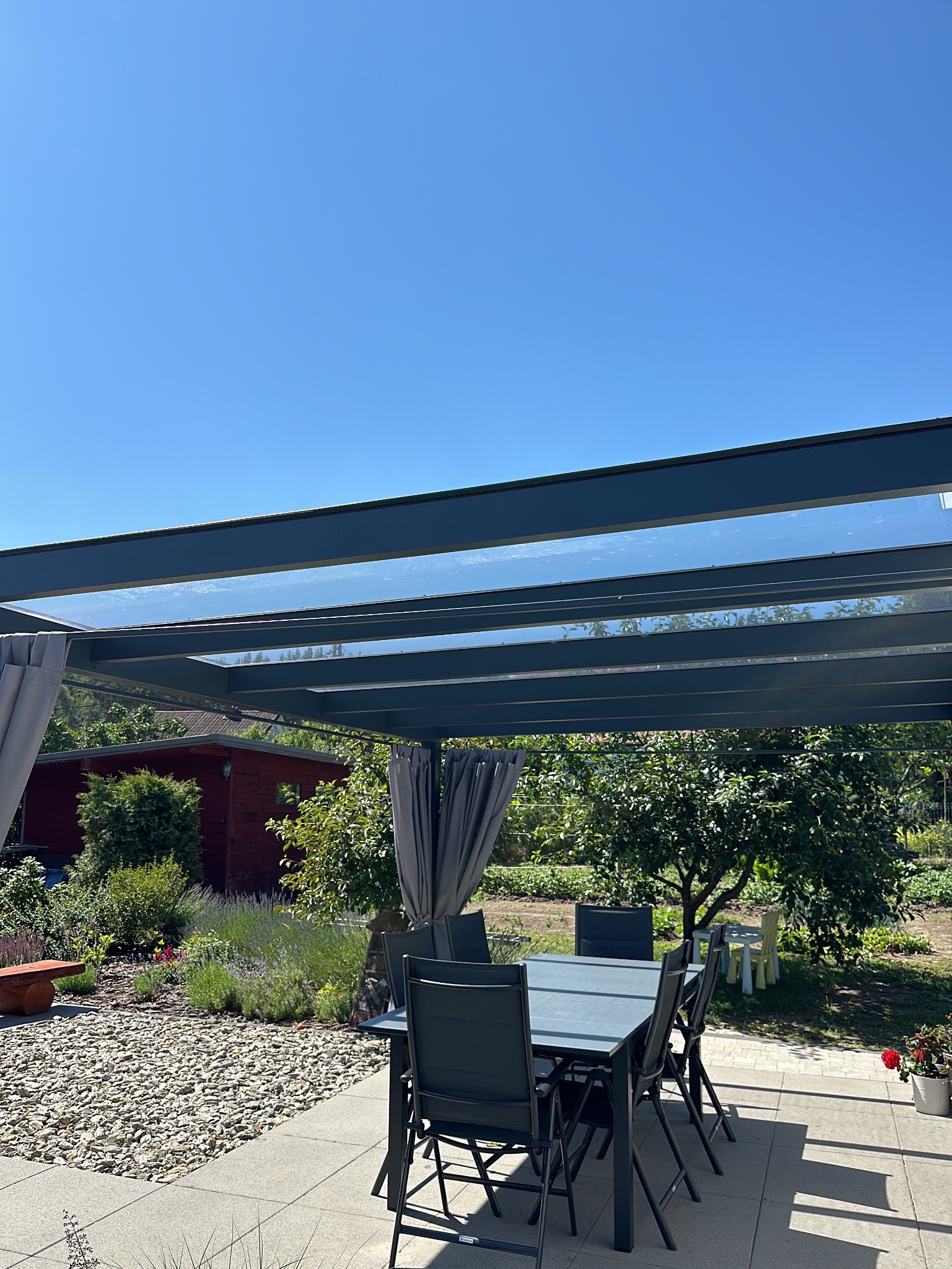 Szklana pergola z metalową konstrukcją i zasłonami na tarasie, w tle ogród z domkiem narzędziowym, widok w słoneczny dzień.