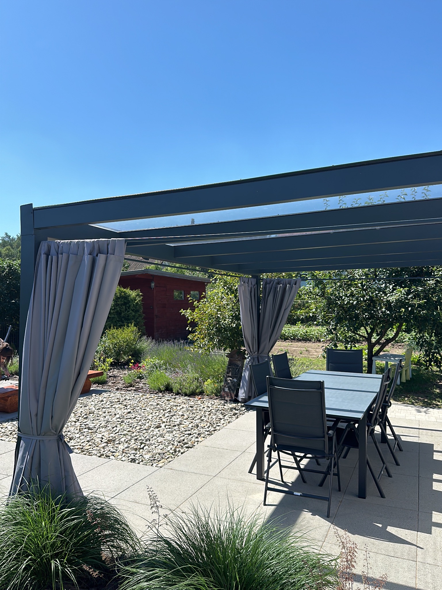 Nowoczesna, antracytowa pergola z zasłonami, szklanym dachem i kompletem mebli ogrodowych na tarasie z jasnych płyt, otoczona zielenią ogrodu i małym drewnianym domkiem.