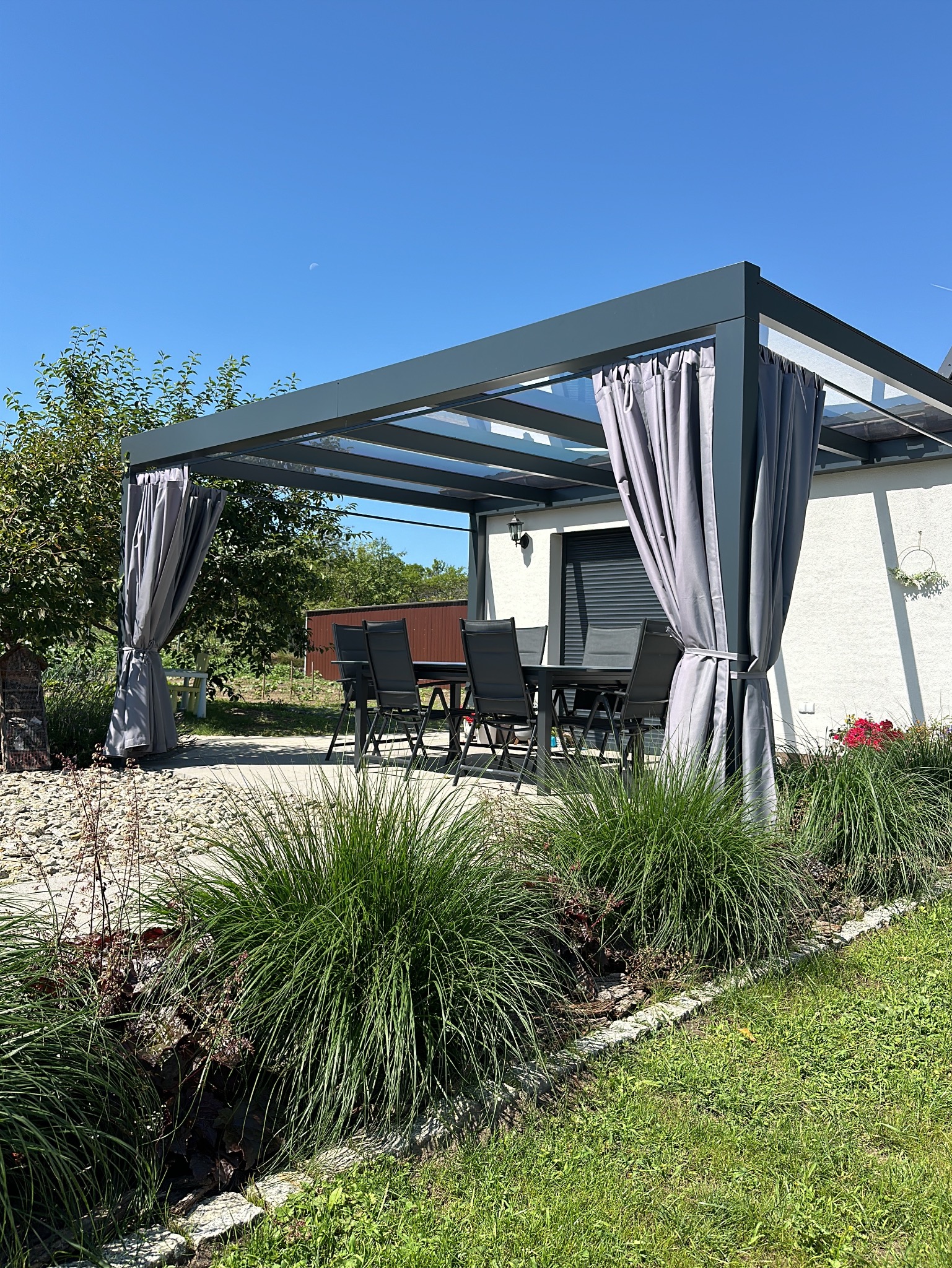 Nowoczesna pergola z szarymi zasłonami i przezroczystym dachem, na tarasie zestaw mebli ogrodowych, w tle biały budynek i błękitne niebo.