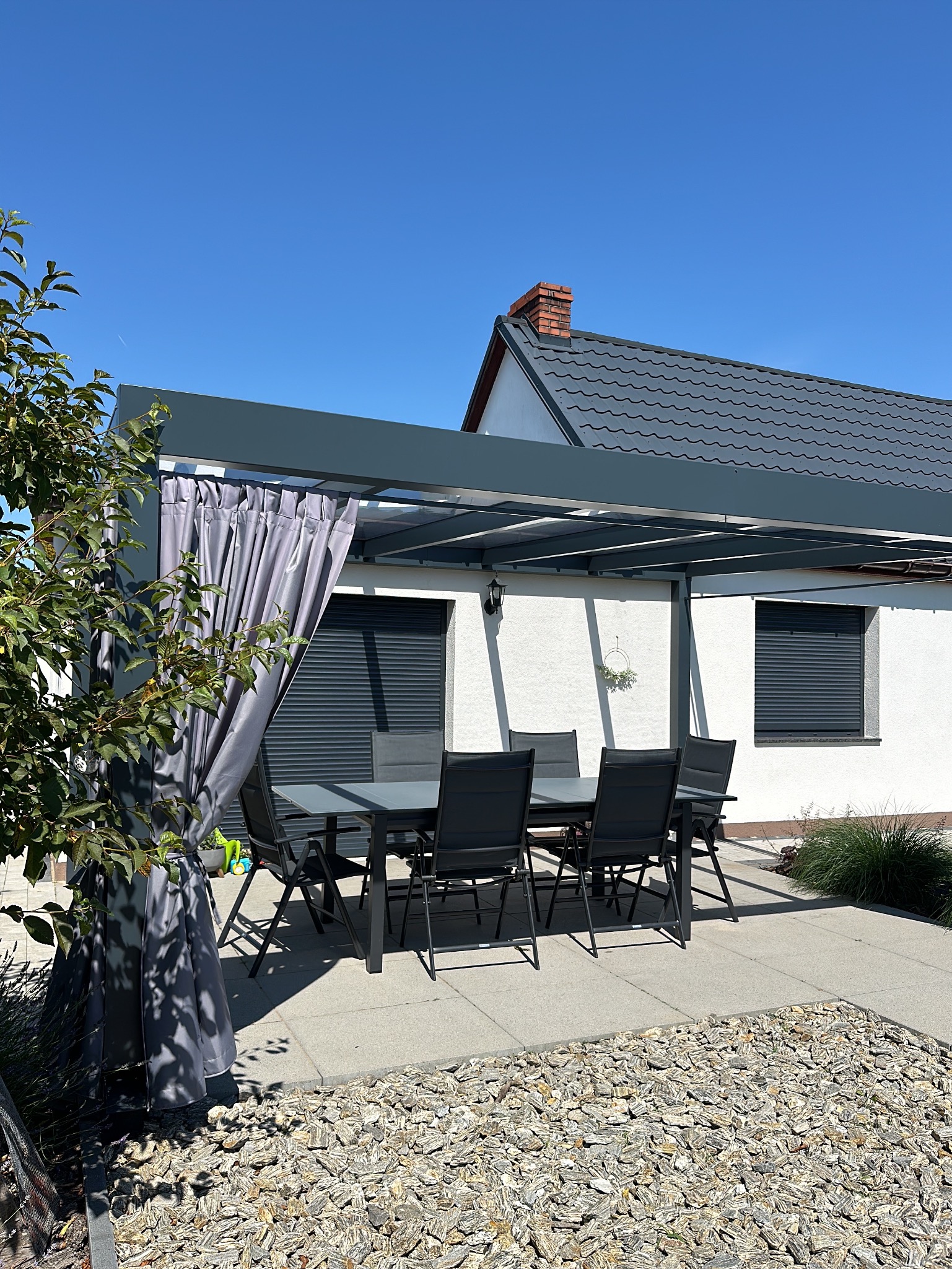 Nowoczesna pergola z szarym zadaszeniem i zasłonami, ustawiona na tarasie z kamienną nawierzchnią, z widokiem na dom z szarą dachówką i roletami.