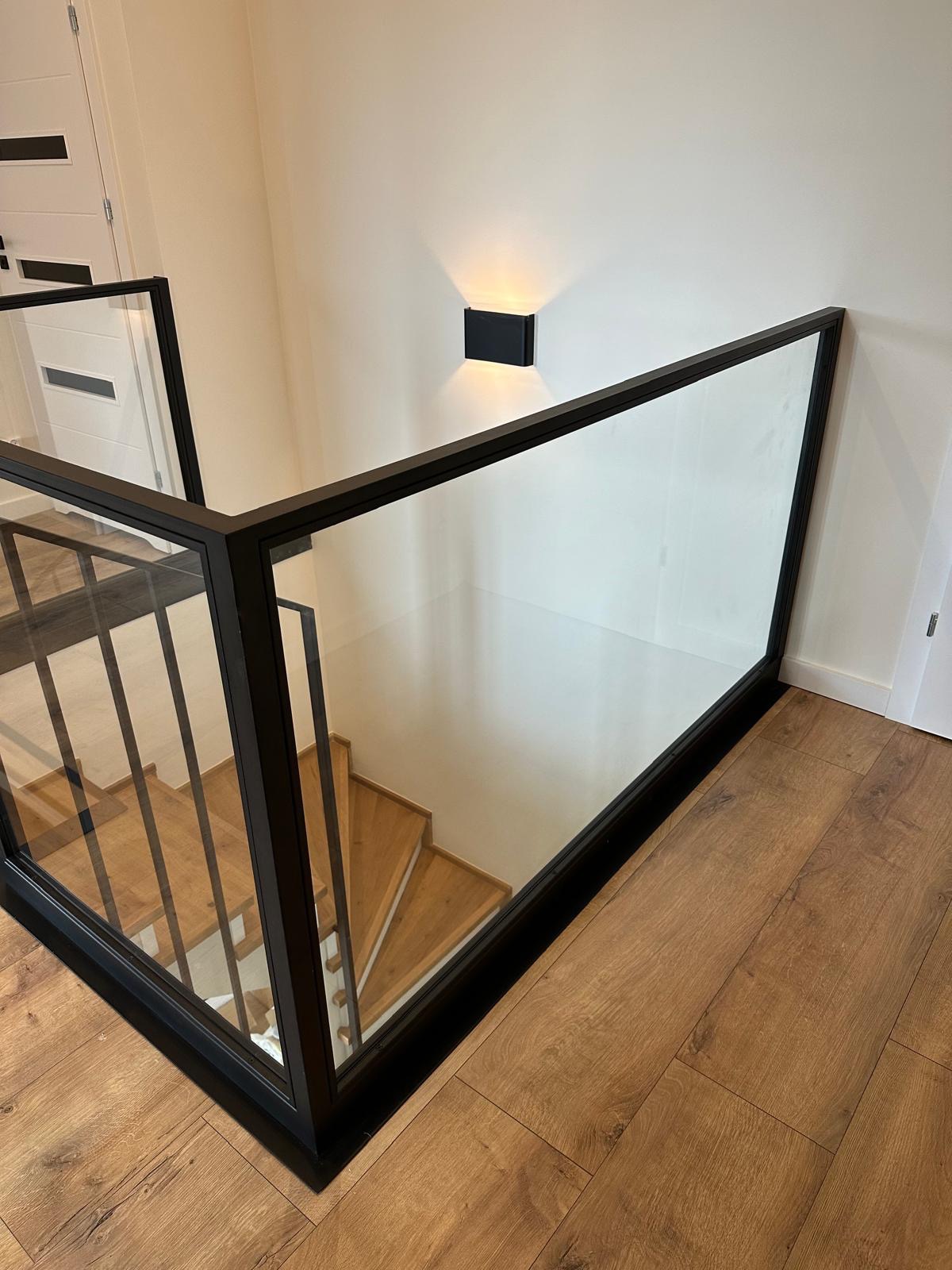 Nowoczesna balustrada szklana z czarną ramą, widok z góry na drewniane schody i minimalistyczny kinkiet. Jasne wnętrze, parkiet w odcieniach brązu.