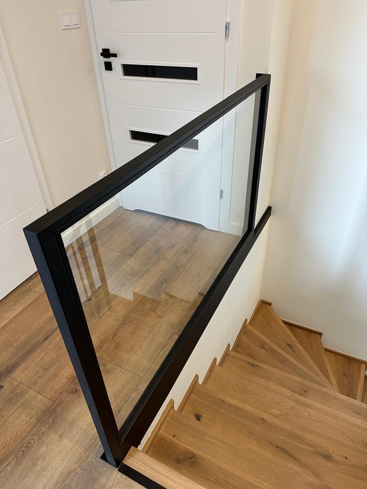 Szklana balustrada schodowa z czarną ramą, widok z góry na drewniane stopnie. Nowoczesny design, jasne wnętrze, drzwi w tle. Minimalistyczny styl.